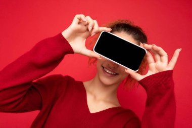 Gülümseyen mutlu, koyu kırmızı süveter giyen, kırmızı arka planda izole edilmiş akıllı telefonlu, cep telefonu ekranlı, boş bir cep telefonu ekranlı, eğlenmek için poz veren genç bir kadının fotoğrafı.