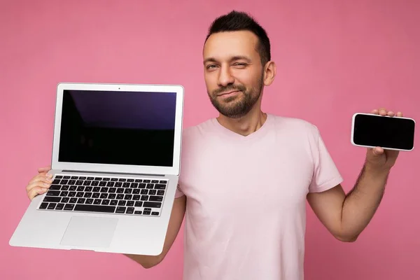 Bonito morena homem segurando computador portátil e telefone celular olhando para a câmera em t-shirt no fundo rosa isolado — Fotografia de Stock