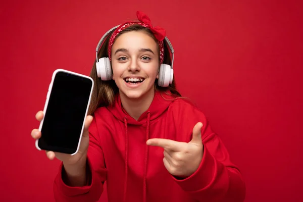 빨간 후드를 착용하고 빨간 배경에서 고립된 웃는 귀여운 갈색 머리 소녀의 사진, 그리고 흰색 무선 헤드폰을 듣기 위해 빈 디스플레이가 있는 스마트폰을 보여 주는 사진. — 스톡 사진