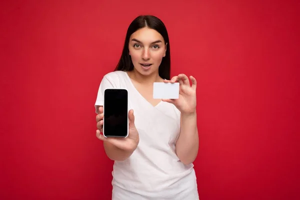 Foto de atraente jovem morena feliz vestindo camiseta branca casual isolada sobre fundo vermelho com espaço vazio segurando na mão telefone celular e mostrando smartphone com tela vazia para — Fotografia de Stock