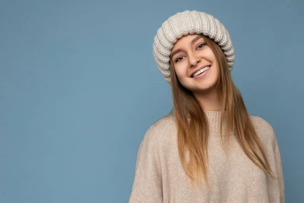 Фото красивой счастливой улыбчивой молодой темноволосой блондинки, изолированной на синем фоне на стене в бежевом тёплом свитере и вязаной бежевой шляпе, смотрящей в камеру — стоковое фото