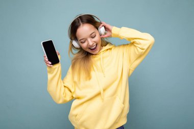 Güzel, neşeli, güler yüzlü, şık kıyafetler giyen, arka plan duvarından izole edilmiş, cep telefonu kullanan, beyaz bluetooth kulaklık takan ve müzik dinleyen genç bir kadının fotoğrafı.