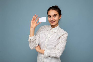 Çekici, pozitif gülümseyen, beyaz bluz giyen esmer kadın elinde kredi kartıyla kameraya bakıyor.