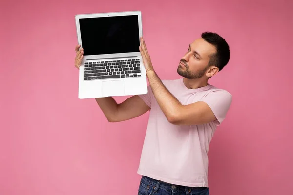 Hombre guapo sosteniendo ordenador portátil mirando monitor portátil en camiseta sobre fondo rosa aislado —  Fotos de Stock