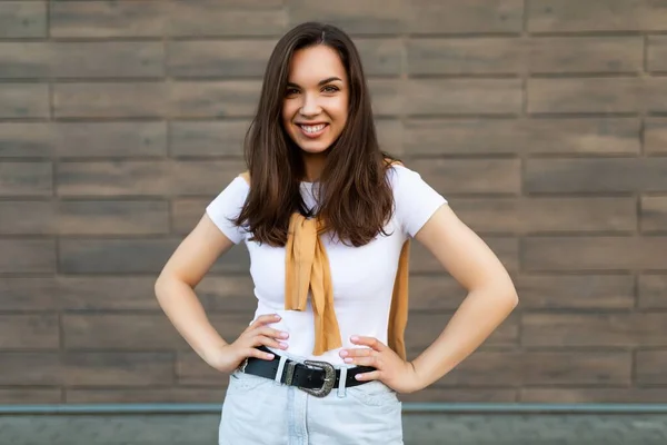 Portrét úspěšné usměvavé radostné mladé brunetky žena na sobě ležérní bílé tričko a džíny se žlutým svetrem balancující v blízkosti hnědé zdi na ulici a baví — Stock fotografie