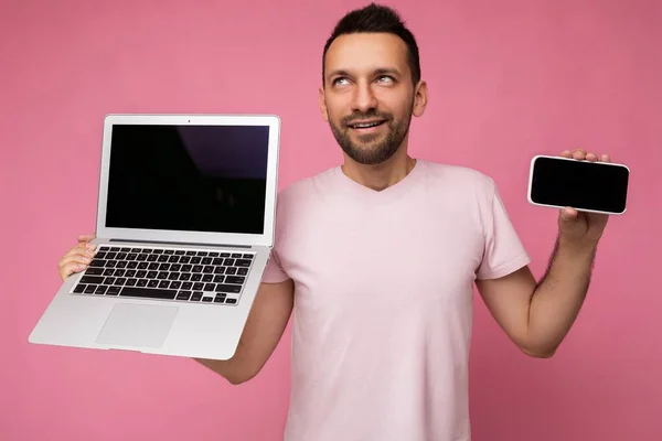 Bonito homem segurando computador portátil e telefone celular olhando para cima em t-shirt no fundo rosa isolado — Fotografia de Stock