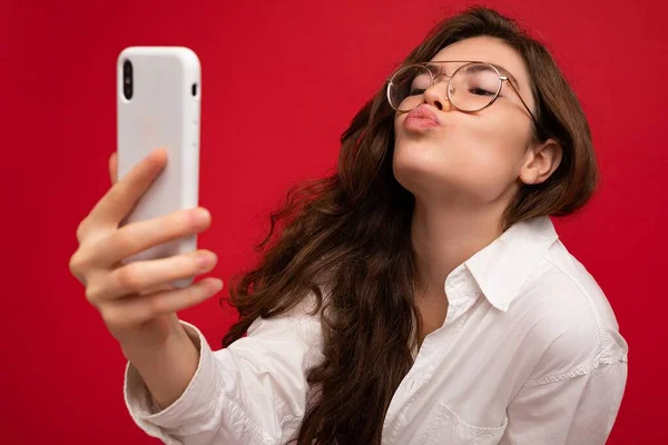 Closeup photo of sexy beautiful positive young brunette woman wearing white shirt and optical glasses isolated over red background holding in hand and using mobile phone taking selfiie looking at — Stock Photo, Image