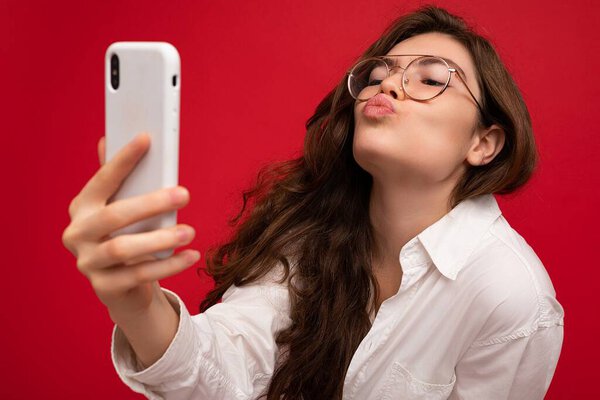 Closeup photo of sexy beautiful positive young brunette woman wearing white shirt and optical glasses isolated over red background holding in hand and using mobile phone taking selfiie looking at