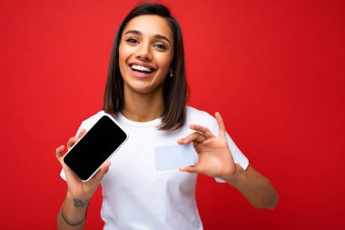 Kırmızı arka plan duvarına izole edilmiş günlük şık beyaz tişört giyen mutlu esmer bir kadının portre fotoğrafı. Model ve kredi kartı yapımı için boş ekranlı telefonu tutuyor.