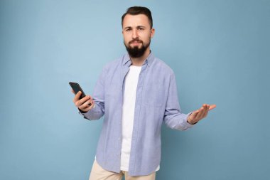 Yakışıklı, üzgün, sakalsız, sakallı, şık beyaz tişörtlü, mavi gömlekli, mavi arka planda, elinde boş bir alan olan genç bir adam.