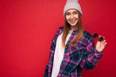 Çekici gülümseyen genç, esmer bir kadının fotoğrafı. Kırmızı arka planda izole edilmiş. Hipster renkli, şık gömlekli, gri şapkalı ve modern güneş gözlüklü bir kadın.