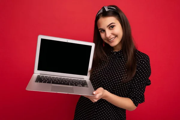 Fénykép gyönyörű mosolygós sötét hajú lány visel fekete ruhát gazdaság netbook nézi kamera elszigetelt színes fal háttér — Stock Fotó