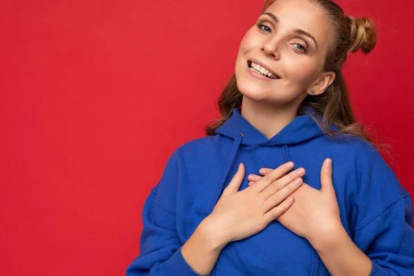 Retrato de jovem feliz positivo sorrindo bela mulher loira com emoções sinceras vestindo casual capuz azul brilhante isolado no fundo vermelho com espaço vazio e de mãos dadas no coração — Fotografia de Stock