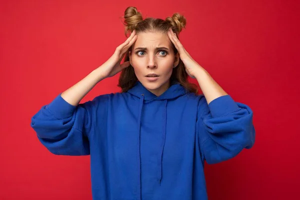 Φωτογραφία της συναισθηματικά σοκαρισμένη νεαρή όμορφη ξανθιά γυναίκα με δύο κέρατα με ειλικρινή συναισθήματα φορώντας hipster φωτεινό μπλε hoodie απομονώνονται σε κόκκινο φόντο με αντίγραφο χώρο — Φωτογραφία Αρχείου