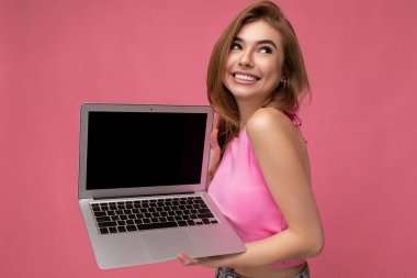 Yan profil fotoğrafı Happy gülümsüyor güzel sarışın kadın bilgisayarında dizüstü bilgisayar tutuyor pembe ekin bluzu giyiyor pembe arka planda izole edilmiş tarafa bakıyor..