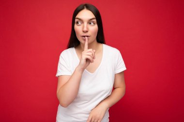 Kırmızı arka planda izole edilmiş ve sessiz kalmak isteyen samimi duyguları olan, samimi beyaz tişörtlü, çekici bir kadının portresi. Şşş jest
