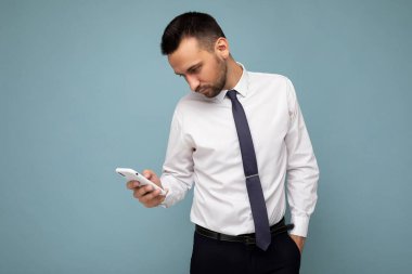 Güzel görünümlü, sakallı, beyaz gömlekli ve kravatlı, mavi arka planda izole edilmiş, el telefonuyla akıllı telefondan iletişim kuran yakışıklı bir esmer.