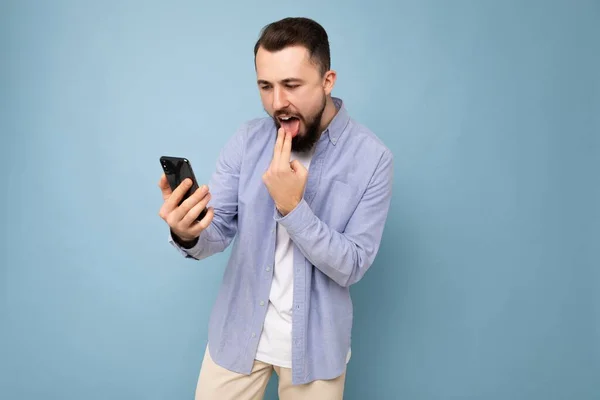 Фото рассерженного красивого бородатого молодого человека в непринужденной стильной одежде, стоящего на заднем плане, с пустым местом в руках и с помощью мобильного телефона, смотрящего смс — стоковое фото