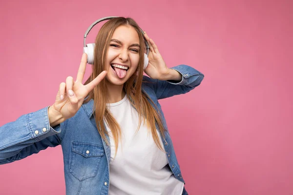 Photo of beautiful positive smiling young blonde woman wearing blue jean shirt and white t-shirt isolated over pink background wearing white wireless bluetooth headphones listening to cool music and — Stock Photo, Image