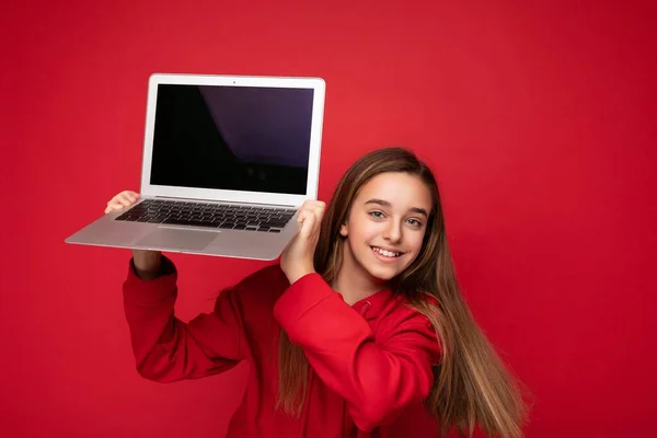 Foto de retrato de cerca de la hermosa chica sonriente feliz con el pelo largo con capucha roja que sostiene el ordenador portátil mirando a la cámara aislada sobre el fondo de la pared roja —  Fotos de Stock