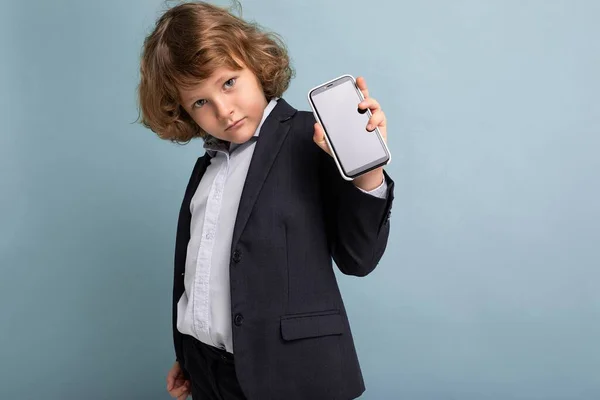 Schöner positiver ernster Junge mit lockigem Haar und Anzug, der das Telefon isoliert über blauem Hintergrund hält, in die Kamera schaut und Smartphone mit leerem Bildschirm zeigt — Stockfoto