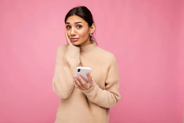 Vonzó aranyos fiatal barna nő visel bézs meleg pulóver áll elszigetelt felett rózsaszín háttér szörfözés az interneten telefonon keresztül nézi a kamera és kétségei vannak — Stock Fotó