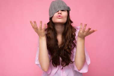 Güzel, duygusal, esmer bir kadının pembe arka plan duvarında dikilip pembe gömlekli, gri şapkalı ve eğlenirken çekilmiş bir fotoğrafı.