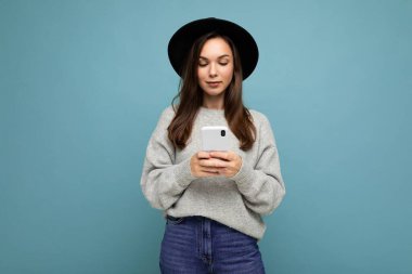 Siyah şapkalı, gri kazaklı güzel bir genç kadın elinde akıllı bir telefonla arka planda izole edilmiş telefon mesajlarına bakıyor.