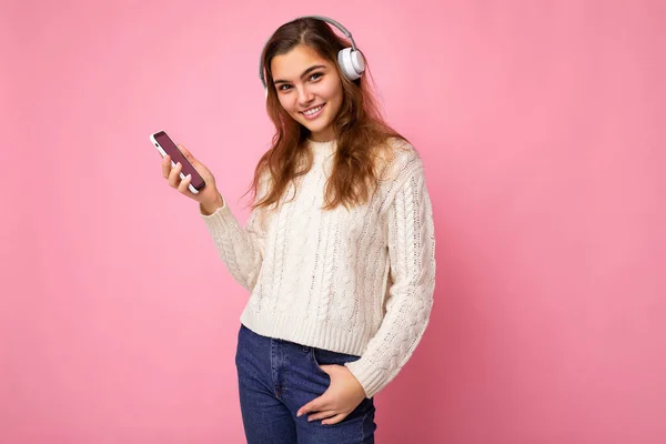 Foto de hermosa mujer joven atractiva positiva usando suéter elegante blanco claro aislado sobre fondo rosa usando auriculares inalámbricos bluetooth blancos y escuchando música fresca y usando el teléfono — Foto de Stock