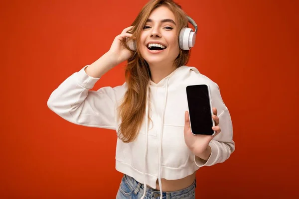 Beautiful happy smiling young woman wearing stylish casual outfit isolated on background wall holding and showing mobile phone with empty display for mockup wearing white bluetooth headphones — Stock Photo, Image