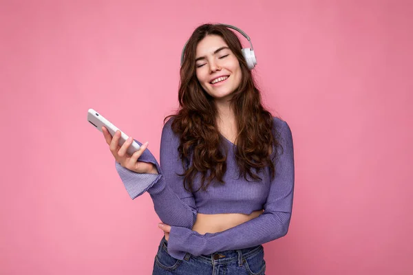 Foto de hermosa mujer joven alegre sonriente con ropa casual elegante aislada sobre la pared de fondo sosteniendo y utilizando el teléfono móvil con auriculares bluetooth blancos escuchando música y — Foto de Stock