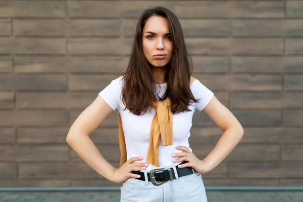 Portrét nedůtklivé smutný uražený uražený rozmrzelý mladý brunet žena na sobě ležérní bílé tričko a džíny se žlutým svetrem balancování u hnědé zdi na ulici — Stock fotografie