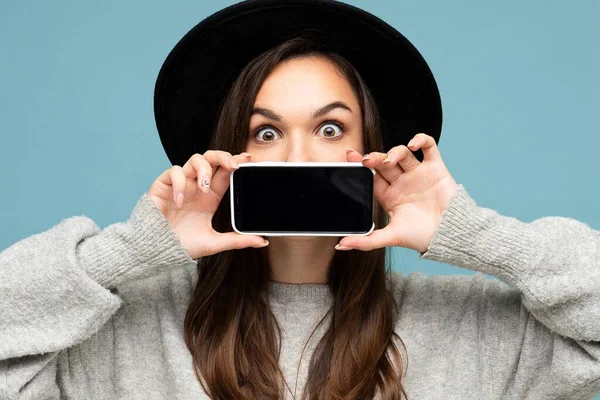 Mulher sorridente muito jovem com belos olhos vestindo chapéu preto e camisola cinza segurando telefone olhando para câmera isolada no fundo — Fotografia de Stock