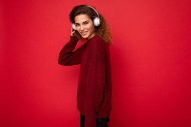Kırmızı arka plan duvarında izole edilmiş koyu kırmızı kazak giyen, beyaz bluetooth kulaklık takan, havalı müzik dinleyen ve eğlenen çekici, pozitif esmer bir kadının yan profil fotoğrafı.