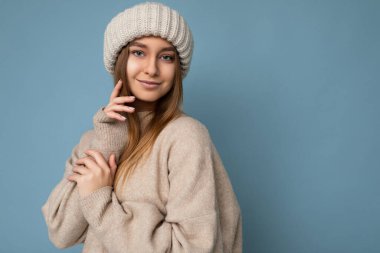 Çekici, mutlu, seksi, esmer, sarışın bir kadının, mavi arka plan duvarında izole edilmiş, bej süveter ve kış bej şapkalı, kameraya bakarken çekilmiş bir fotoğrafı.