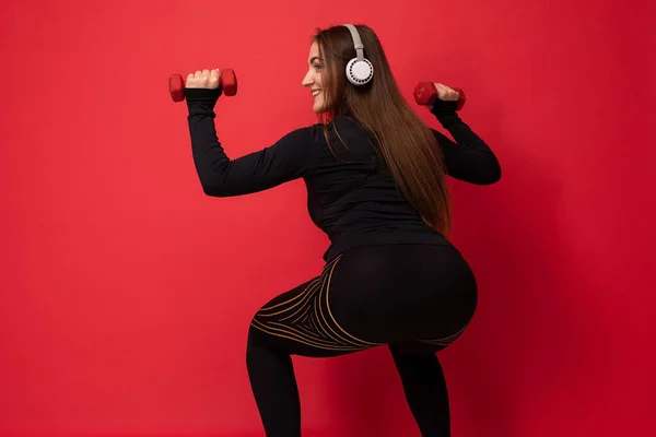 Belle jeune femme brune souriante et heureuse portant des vêtements de sport noirs isolés sur un mur de fond rouge faisant du squat de remise en forme à l'aide d'haltères portant des écouteurs bluetooth blancs écoutant de la musique — Photo