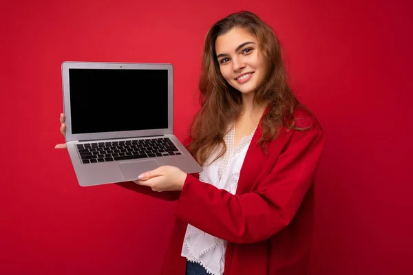 Photo of Beautiful smiling happy young brunette woman holding computer laptop with empty monitor screen wearing red cardigan and white blouse looking at camera isolated over red background — Stock Photo, Image