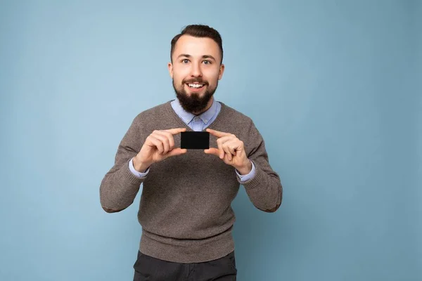 Handsome smiling brunette bearded man wearing grey sweater and blue shirt isolated on background wall holding credit card looking at camera — Stock Photo, Image