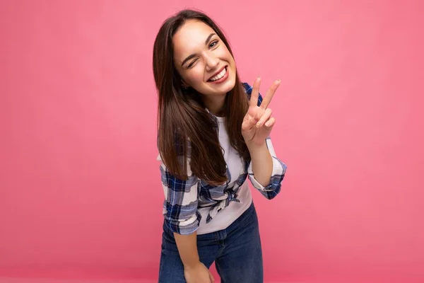 Photo shot of young positive delightful smiling pretty brunette woman with sincere emotions wearing hipster check shirt standing isolated on pink background with free space and showing peace sign — Stock Photo, Image