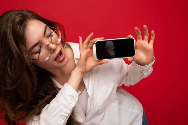 Foto de primer plano de la hermosa mujer morena sexy con camisa blanca y gafas ópticas aisladas sobre fondo rojo sosteniendo en la mano y mostrando el teléfono móvil con pantalla vacía para recortar buscando — Foto de Stock