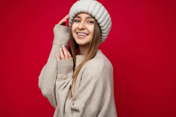 Retrato de foto de sexy lindo atractivo feliz sonriente joven rubia oscura de pie aislado sobre la pared de fondo rojo con suéter beige y sombrero beige mirando a la cámara —  Fotos de Stock