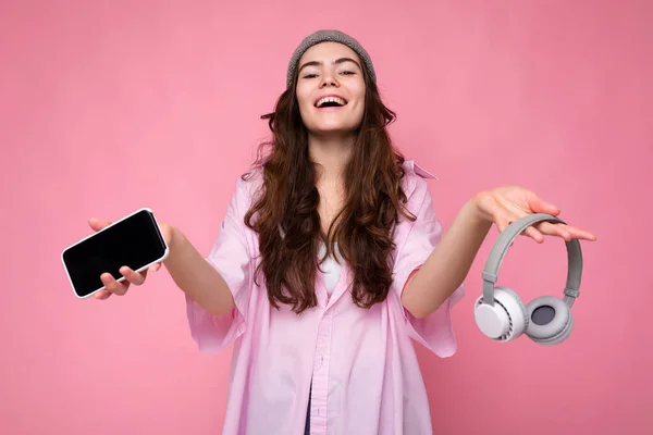 Foto de hermosa mujer joven sonriente positiva con elegante traje casual aislado en la pared de fondo colorido celebración de auriculares inalámbricos bluetooth blanco y mostrando el teléfono móvil con la pantalla vacía — Foto de Stock