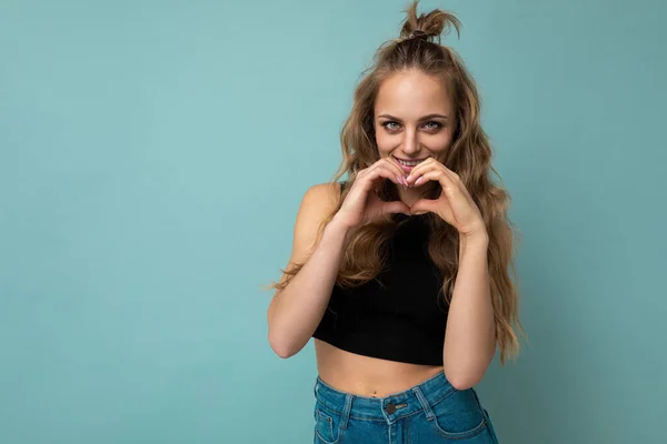 Foto de jovem attarctive mulher loira escura encaracolado com emoções sinceras vestindo top preto isolado sobre fundo azul com espaço vazio e mostrando gesto do coração — Fotografia de Stock