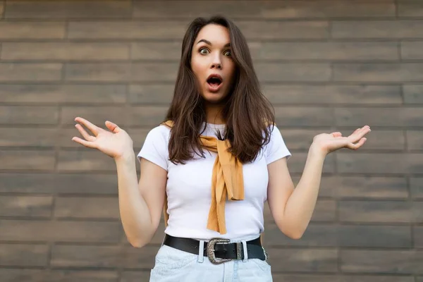 Foto de joven emocionalmente conmocionada mujer morena hermosa asombrada vistiendo ropa casual de pie en la calle cerca de la pared. Concepto de estilo de vida — Foto de Stock