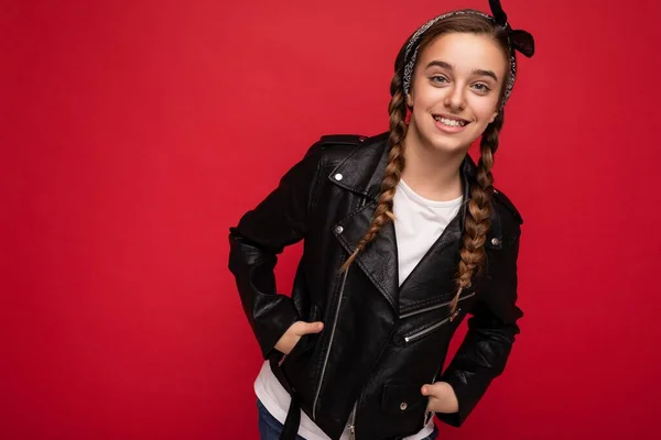 Shot of attractive positive smiling brunette little female teenager with pigtails wearing stylish black leather jacket and white t-shirt isolated over red background wall looking at camera — Stock Photo, Image