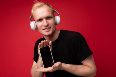 Yakışıklı genç sarışın adam sıradan siyah tişörtler giyiyor ve kulaklık takıyor. Podcast dinliyor. Elinde boş ekranlı akıllı telefon var.