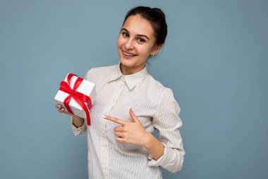 Güzel, mutlu, genç esmer kadın renkli arka plan duvarında izole edilmiş şık günlük kıyafetler giymiş elinde hediye kutusuyla kameraya bakıyor.