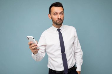 Yakışıklı, güzel, tıraşsız, sakallı, beyaz gömlekli, kravatlı, mavi arka planda izole edilmiş, cep telefonuyla iletişim kuran bir erkek.