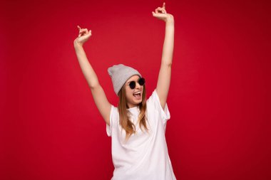 Güzel, pozitif gülümseyen, koyu saçlı, kırmızı arka planda izole edilmiş, gri şapkalı, beyaz tişörtlü güzel bir kadının fotoğrafı. Model ve şık güneş gözlükleri takıyor ve eğleniyor.