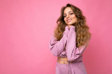 Genç, kıvırcık, seksi ifadeli, neşeli ve mutlu yüzlü pembe spor takım elbiseli, pembe arka planda izole edilmiş, fotokopi alanı olan bir kadın.